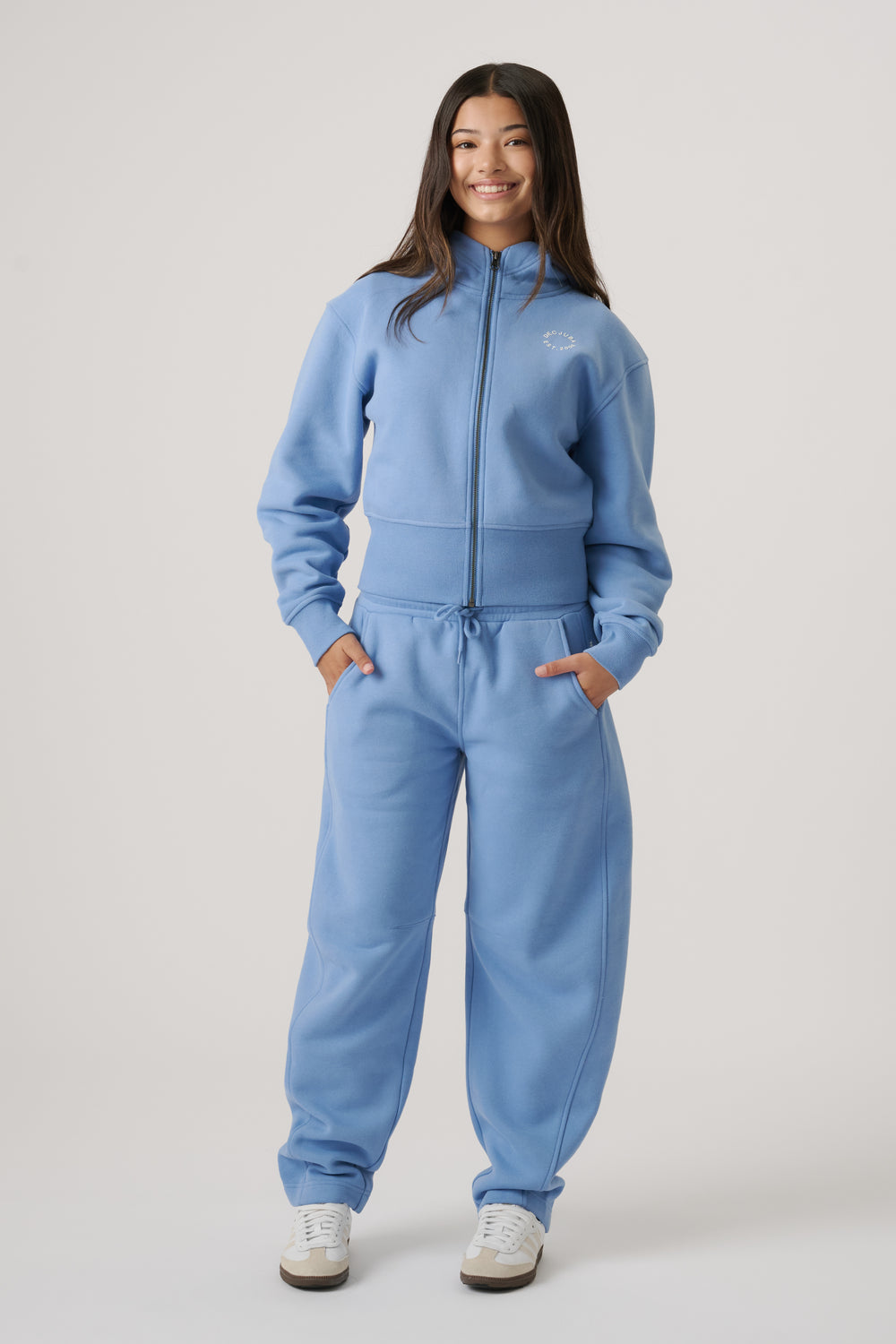 Girl wearing relaxed dusty blue barrel-leg trackpants paired with a matching zip-up jacket and white sneakers.