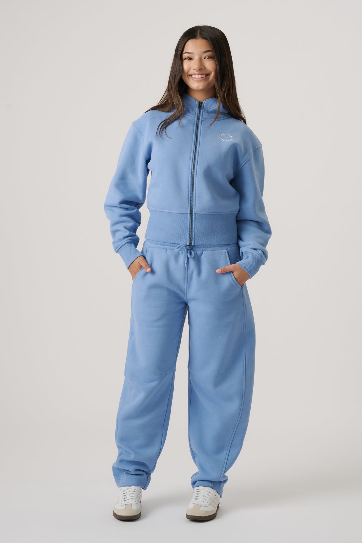 Girl wearing relaxed dusty blue barrel-leg trackpants paired with a matching zip-up jacket and white sneakers.