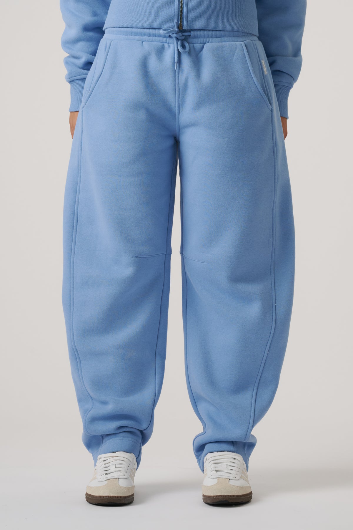 Close-up of relaxed-fit dusty blue barrel leg trackpants with drawstring waist and pockets, styled with sneakers.