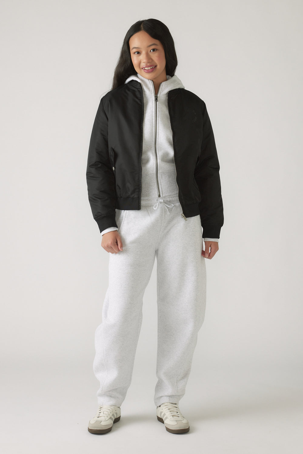Girl wearing light grey barrel leg pants with a grey zip-up hoodie and a black bomber jacket, smiling confidently