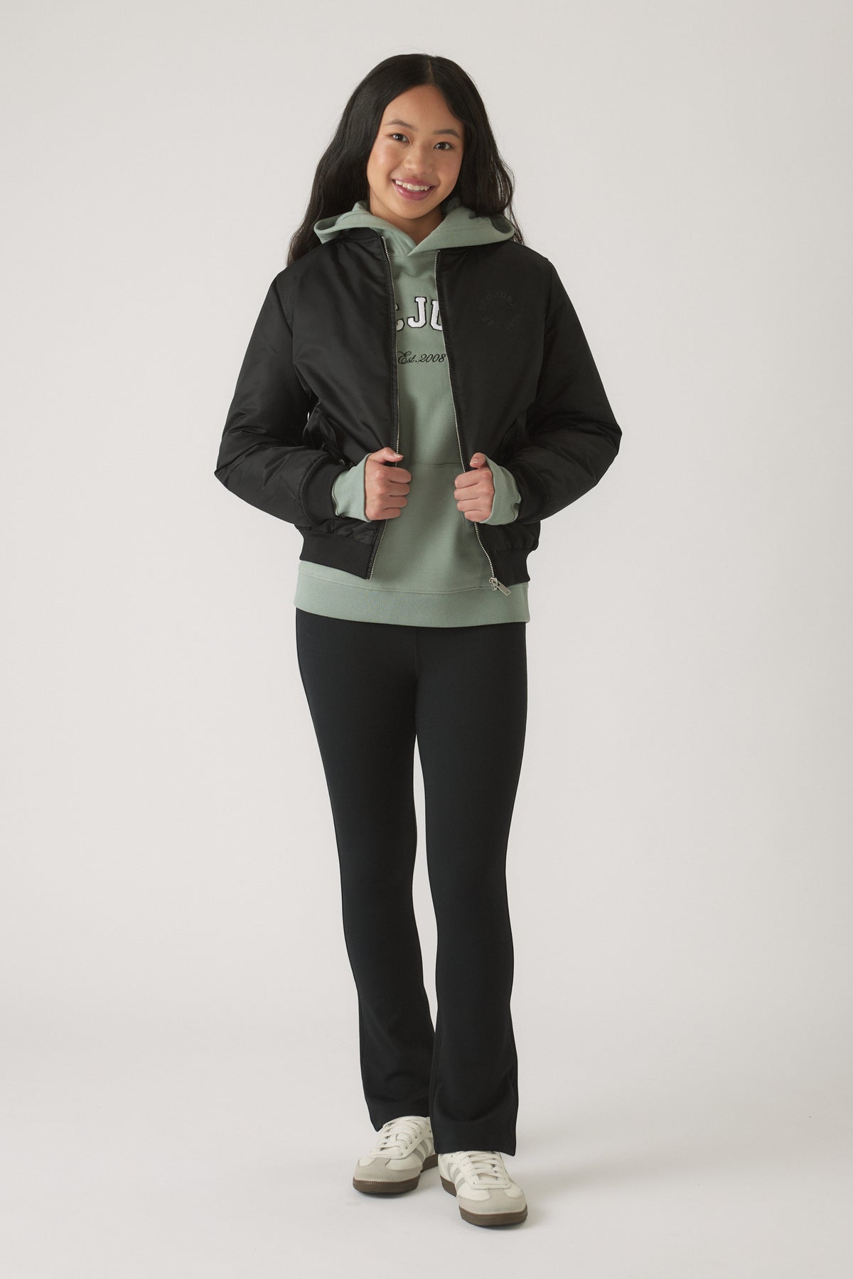 Girl wearing a black bomber jacket layered over a sage green hoodie with black fitted pants and white sneakers smiling