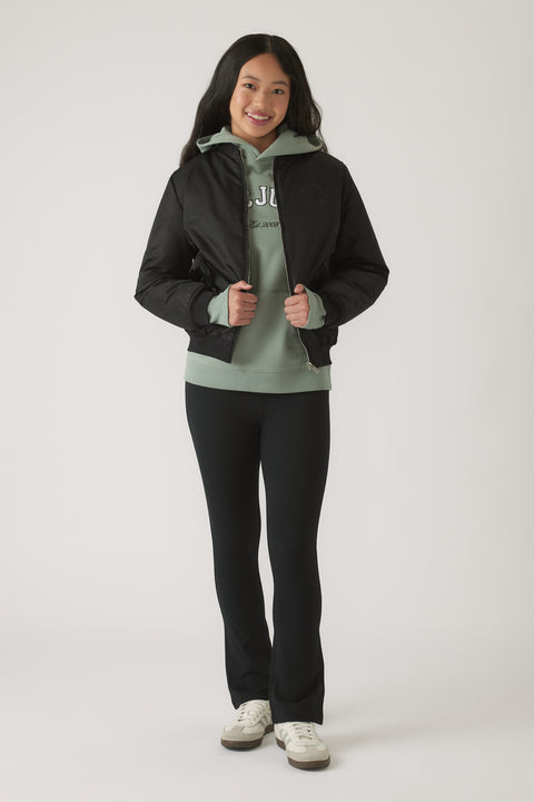 Girl wearing a black bomber jacket layered over a sage green hoodie with black fitted pants and white sneakers smiling