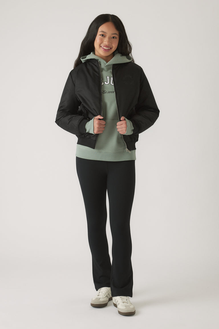 Girl wearing a black bomber jacket layered over a sage green hoodie with black fitted pants and white sneakers smiling
