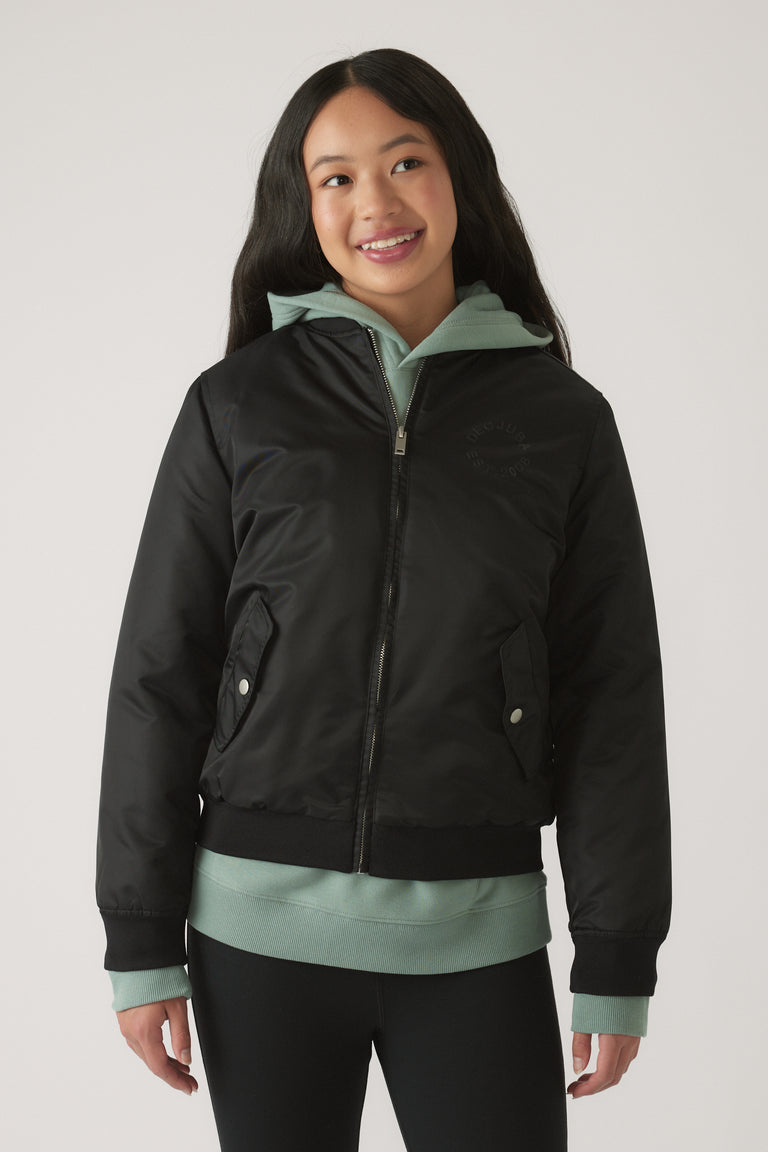 Girl wearing a black nylon bomber jacket layered over a green hoodie with black leggings, smiling and standing casually.