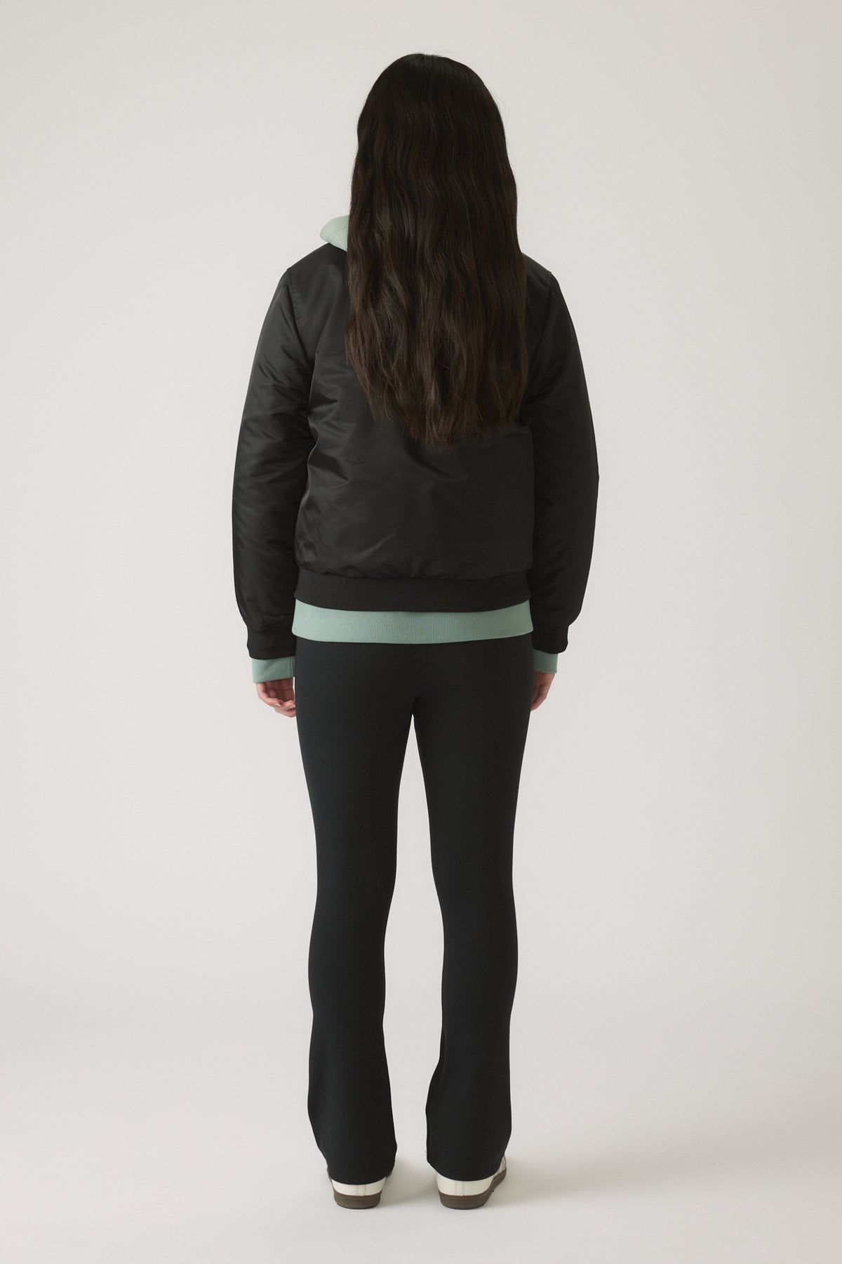 Girl wearing a black nylon bomber jacket layered over a mint green hoodie, paired with slim black pants and white shoes
