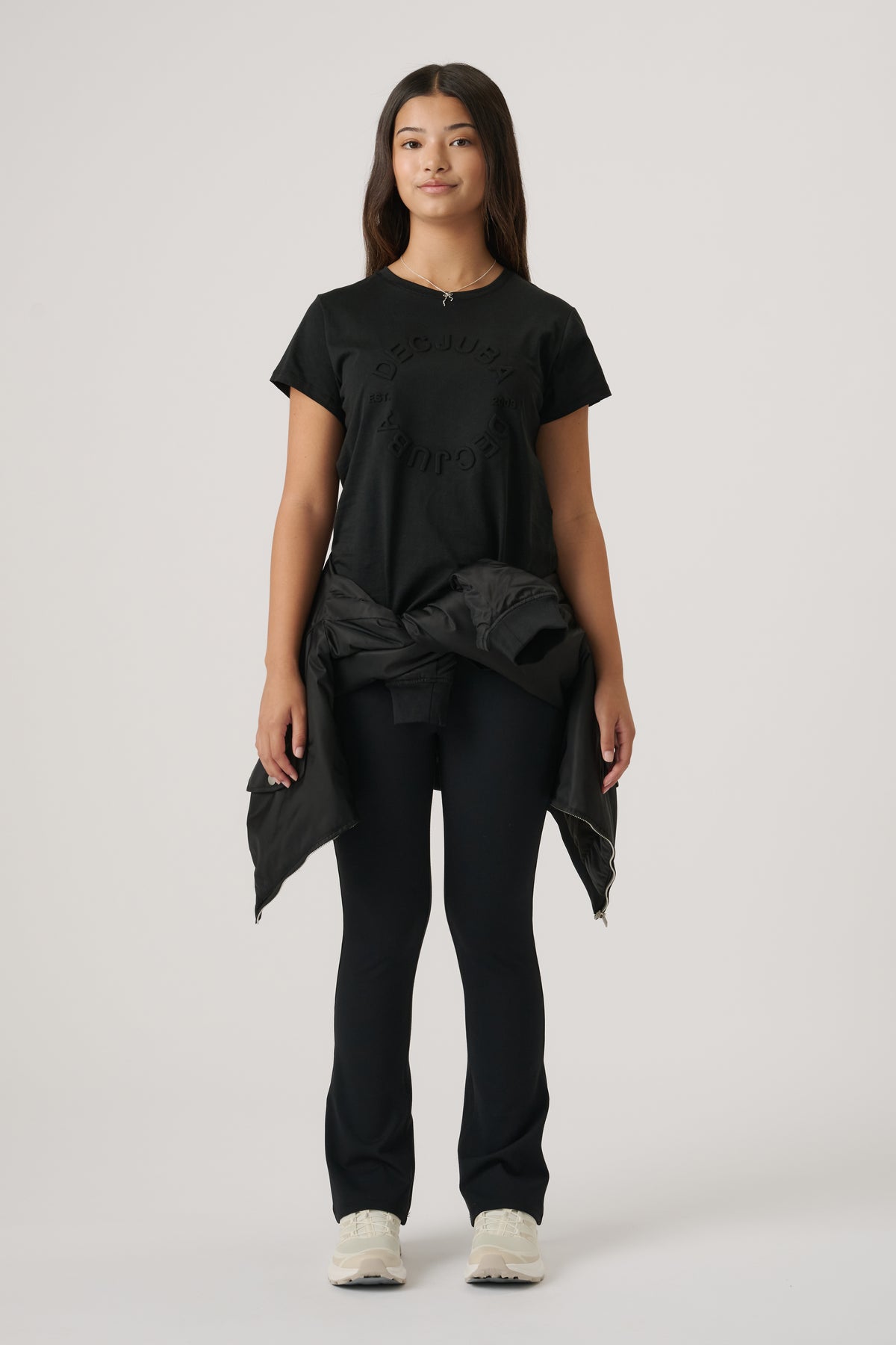 Teen girl wearing a black embossed logo tee paired with black fitted pants and a jacket tied around the waist