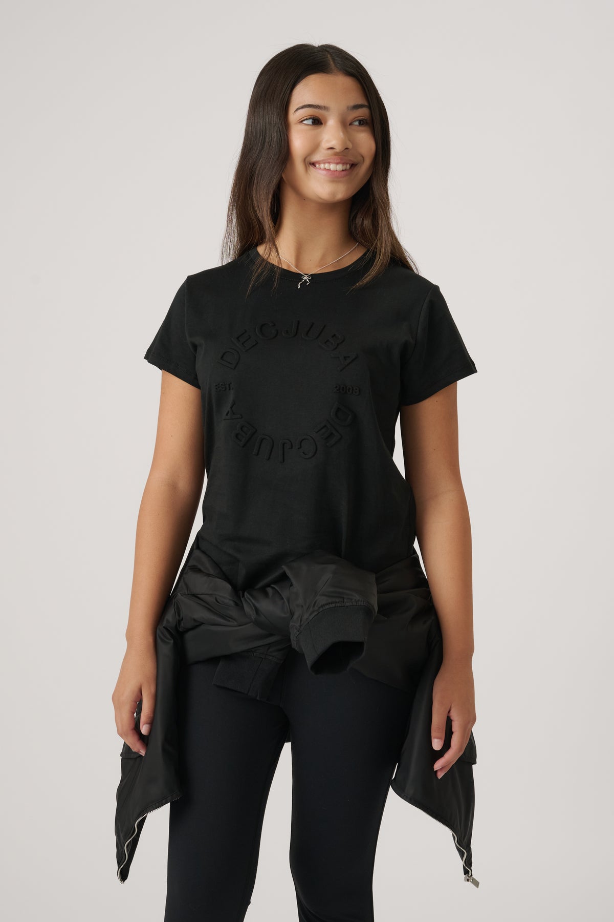 Teen girl smiling wearing a black embossed logo tee with a casual jacket tied around her waist and black leggings
