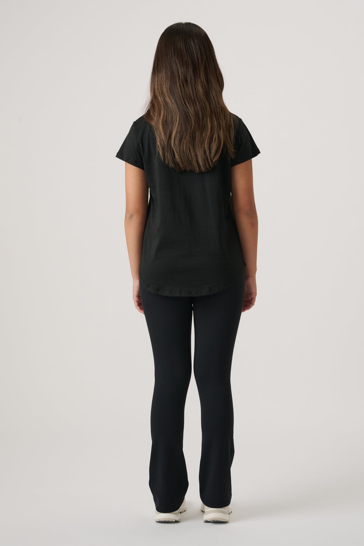 Girl standing with back to camera wearing a fitted black tee and matching slim flare pants with white sneakers.