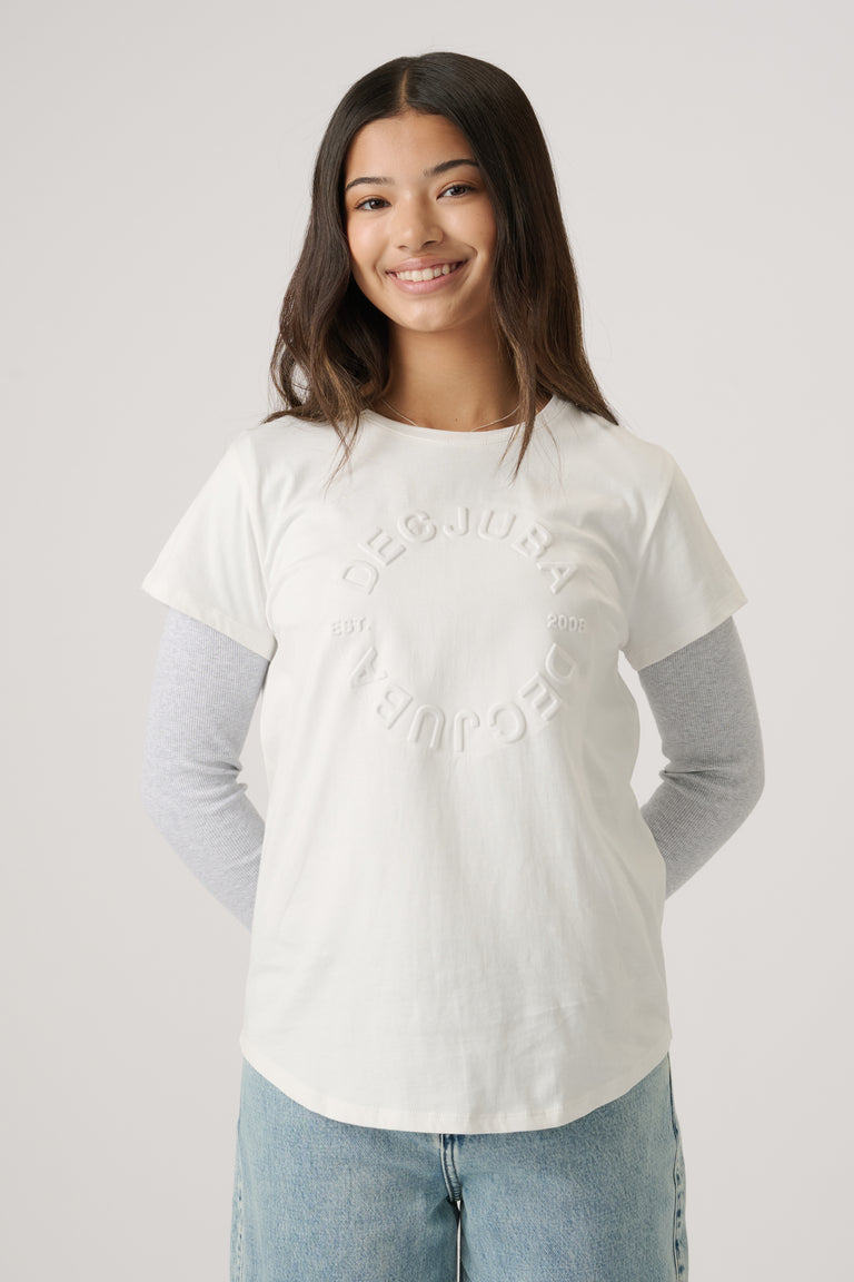 Teen girl smiling wearing a white embossed tee layered over a grey long-sleeve top with light blue jeans.