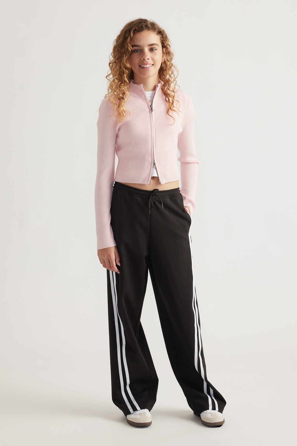 Teen girl wearing a light pink zip-up knit top paired with loose black track pants featuring white side stripes