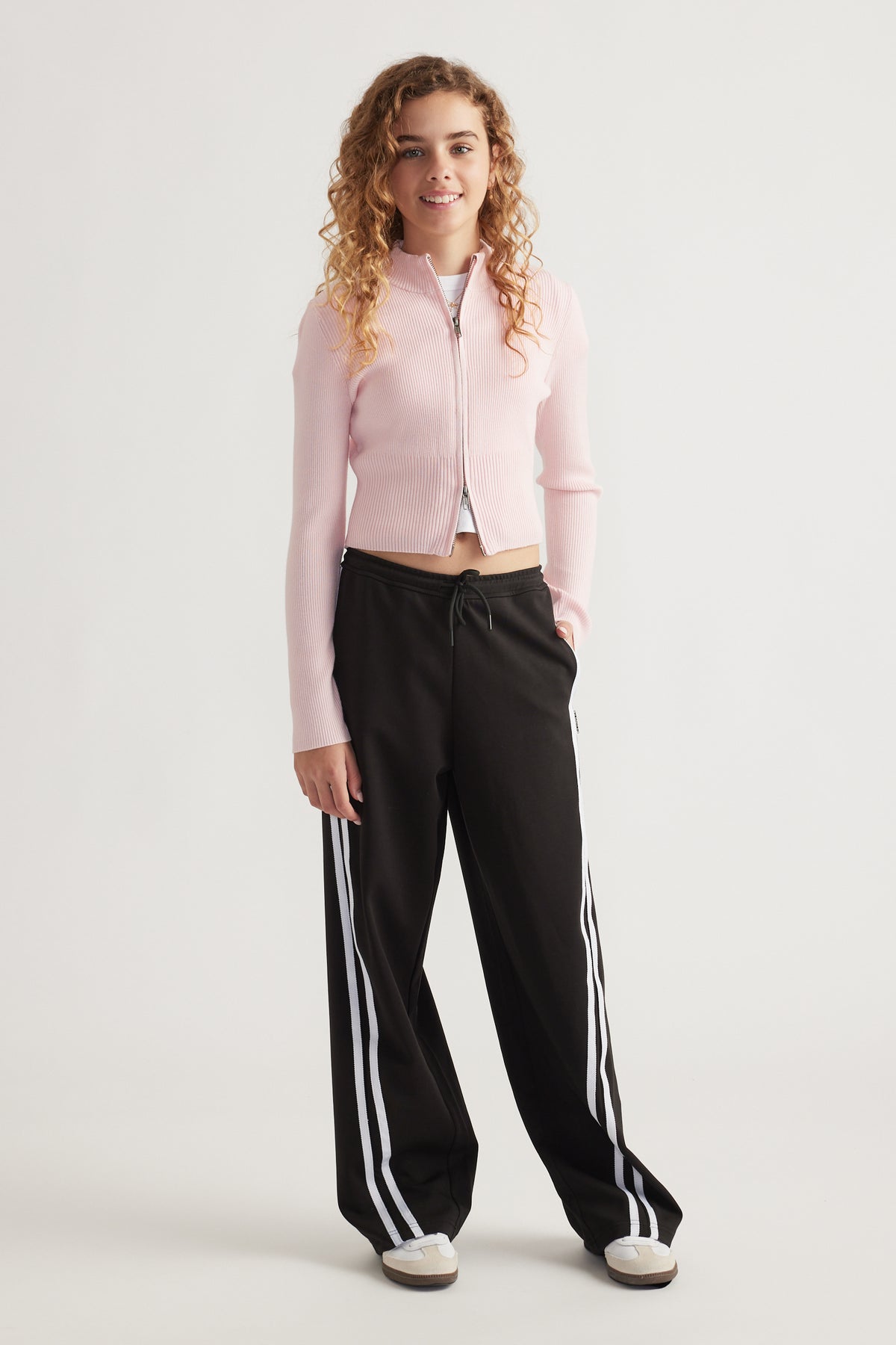 Teen girl wearing a light pink zip-up knit top paired with loose black track pants featuring white side stripes