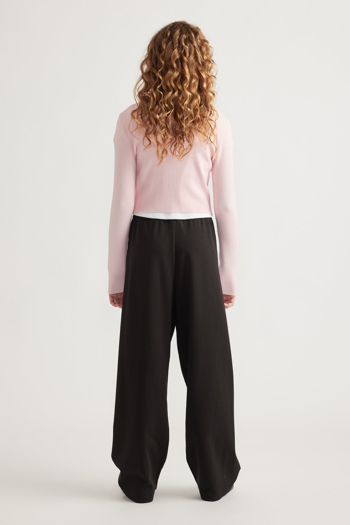Rear view of girl wearing a pale pink zip-up knit with stretch fabric and loose black wide-leg pants