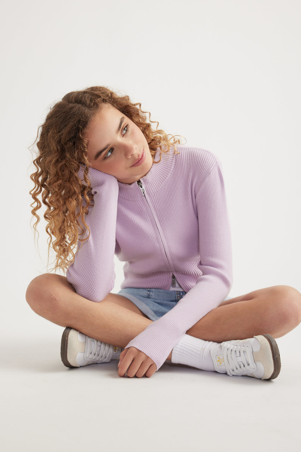 Girl seated in a lilac zip front knit top with a stand collar, denim shorts and white sneakers