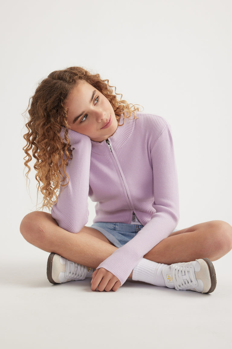 Girl seated in a lilac zip front knit top with a stand collar, denim shorts and white sneakers