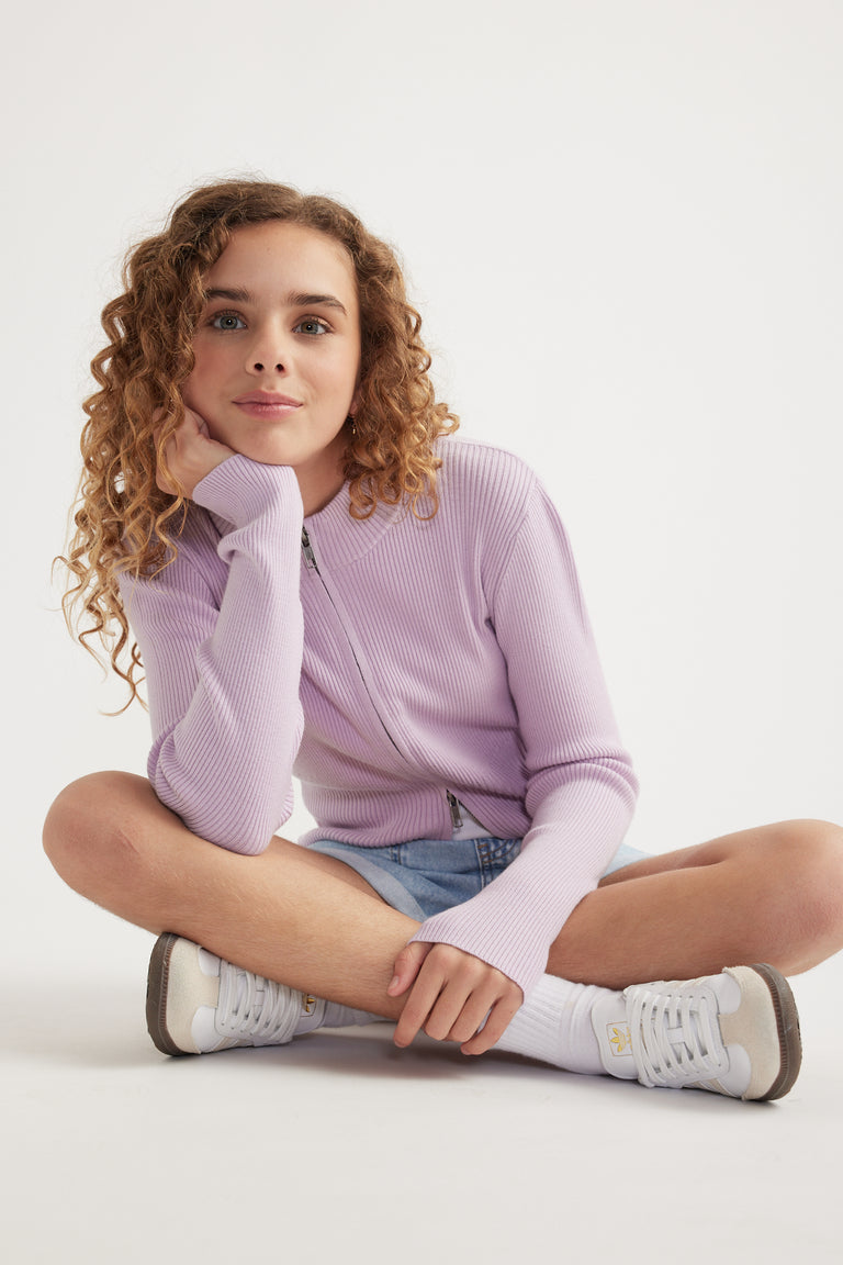Girl sitting cross legged in a lilac zip up knit top, denim shorts and white sneakers