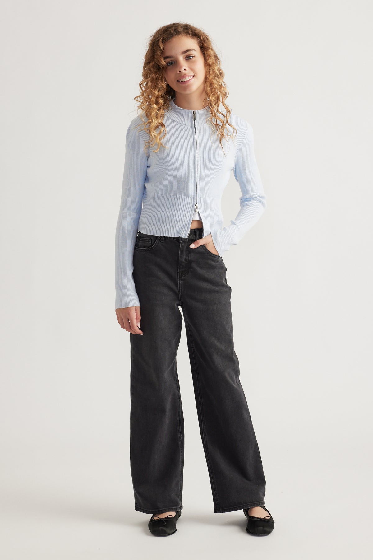 Teen girl smiling wearing a light blue zip-up knit with a stand collar and relaxed black wide-leg jeans