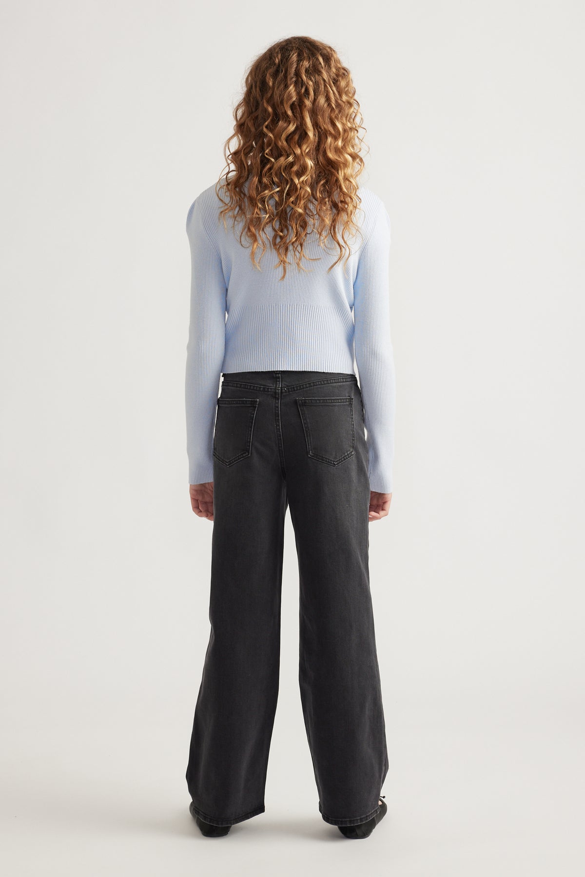 Rear view of girl with curly hair wearing a pale blue fitted knit top paired with loose black wide-leg jeans