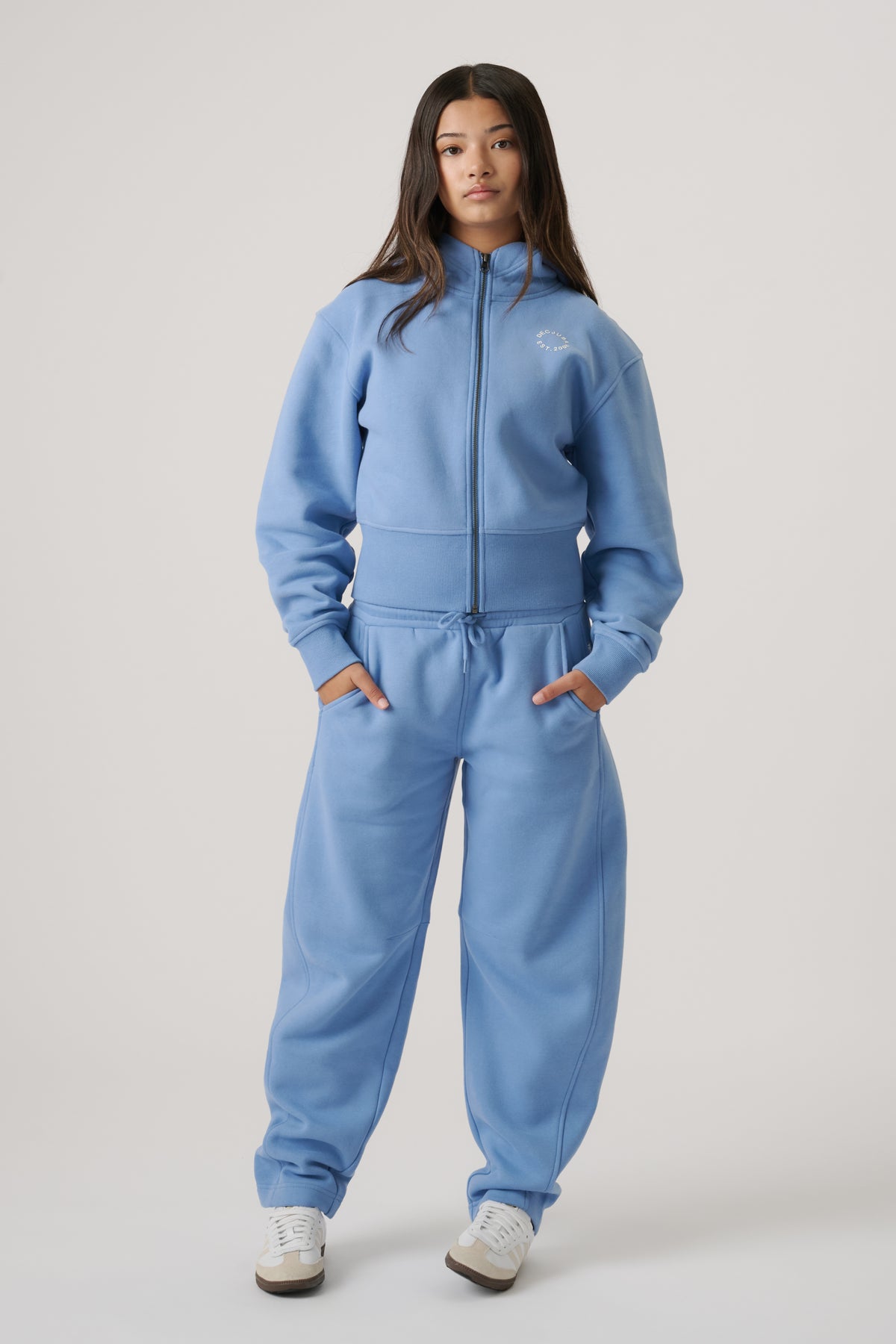 Teen girl wearing a dusty blue cropped zip hoodie and matching joggers, styled casually with white sneakers.