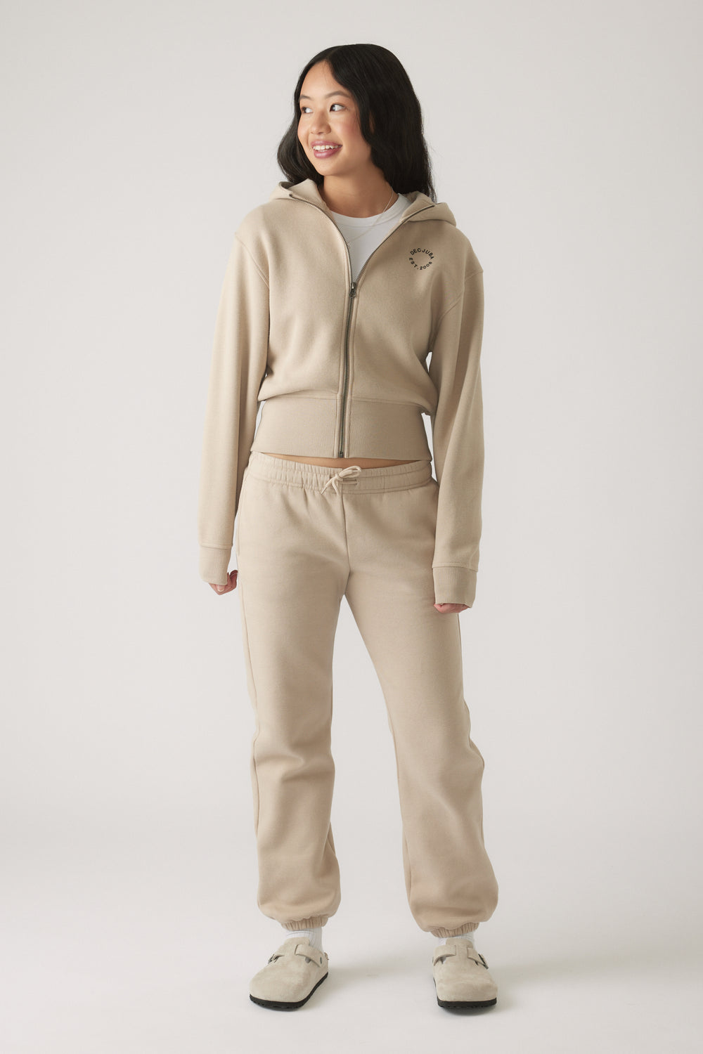 Teen girl wearing a beige cropped zip hoodie with matching drawstring joggers, smiling and looking to the side