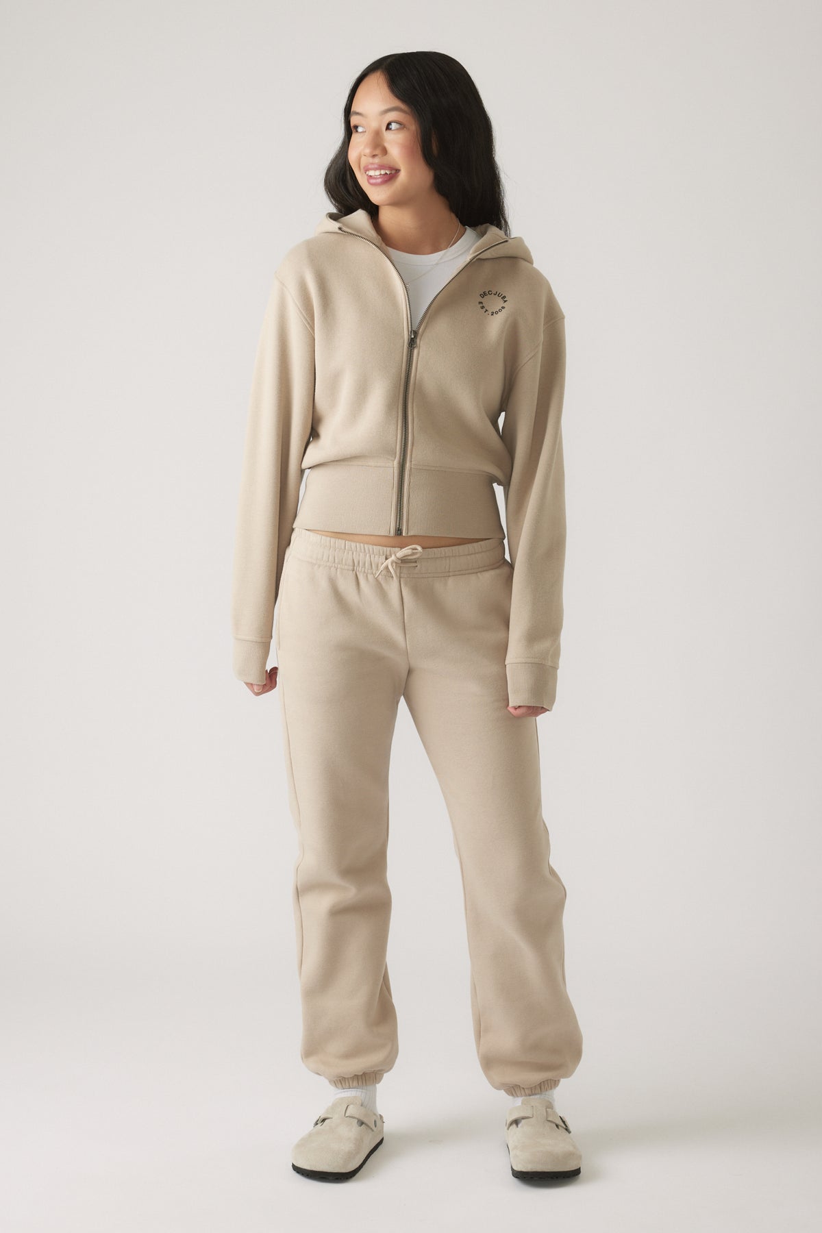 Teen girl wearing a beige cropped zip hoodie with matching drawstring joggers, smiling and looking to the side