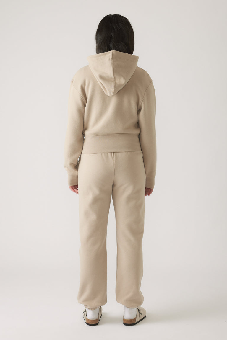 Girl wearing a beige zip-up cropped hoodie and matching joggers, shown from behind with sneakers against a plain background.