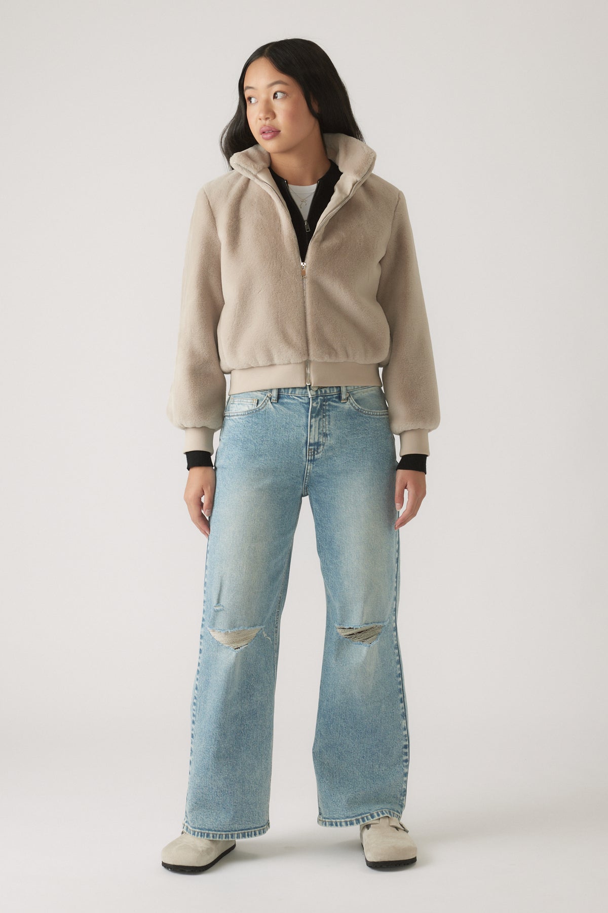 Teen girl wearing light blue baggy ripped jeans paired with a beige zip-up fleece jacket and casual shoes
