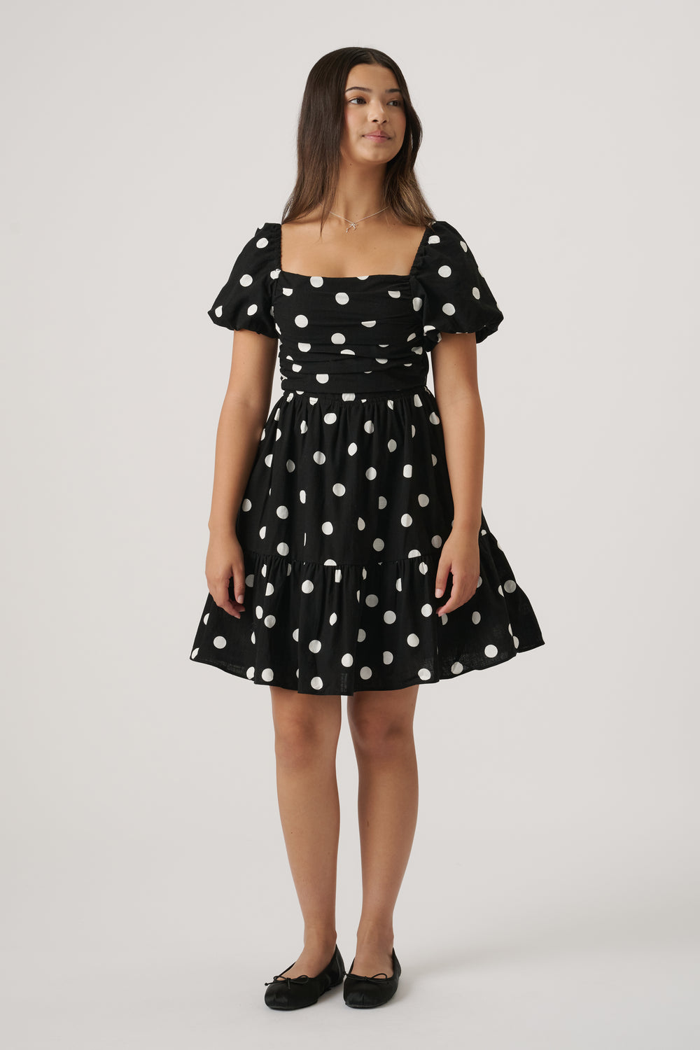 Young girl wearing a black mini dress with white polka dots, puff sleeves, and ballet flats, standing against plain background