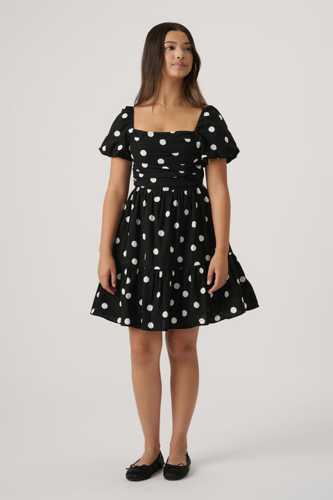Young girl wearing a black mini dress with white polka dots, puff sleeves, and ballet flats, standing against plain background