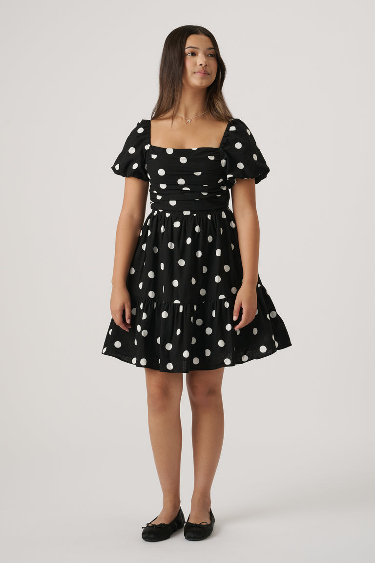 Young girl wearing a black mini dress with white polka dots, puff sleeves, and ballet flats, standing against plain background