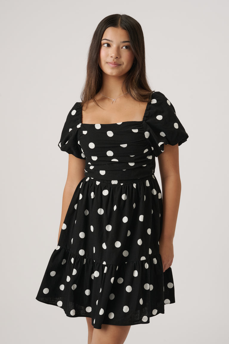 Young woman wearing a black mini dress with white polka dots, puff sleeves, and a ruched bodice standing against a plain background.