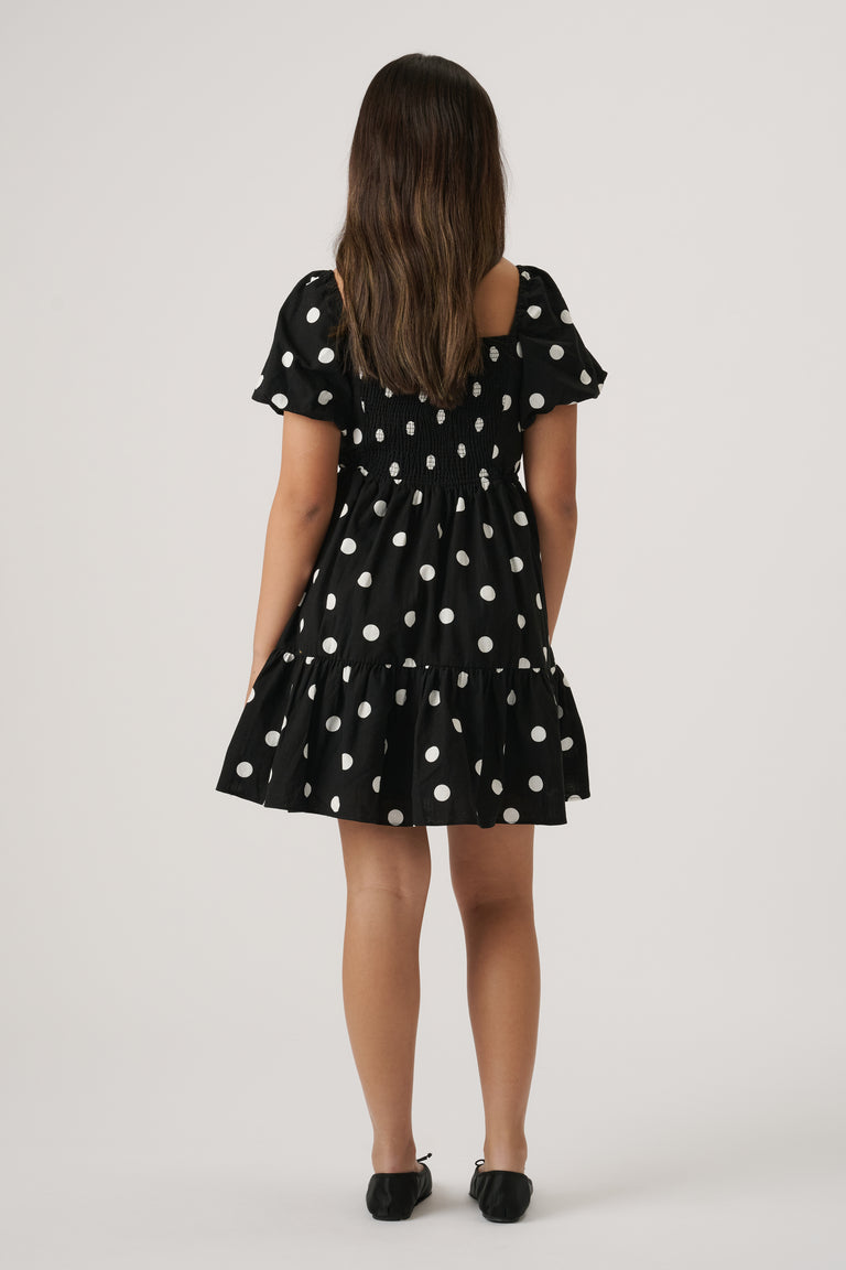 Young girl wearing a black mini dress with white polka dots, puff sleeves, and a gathered tiered skirt, black flats