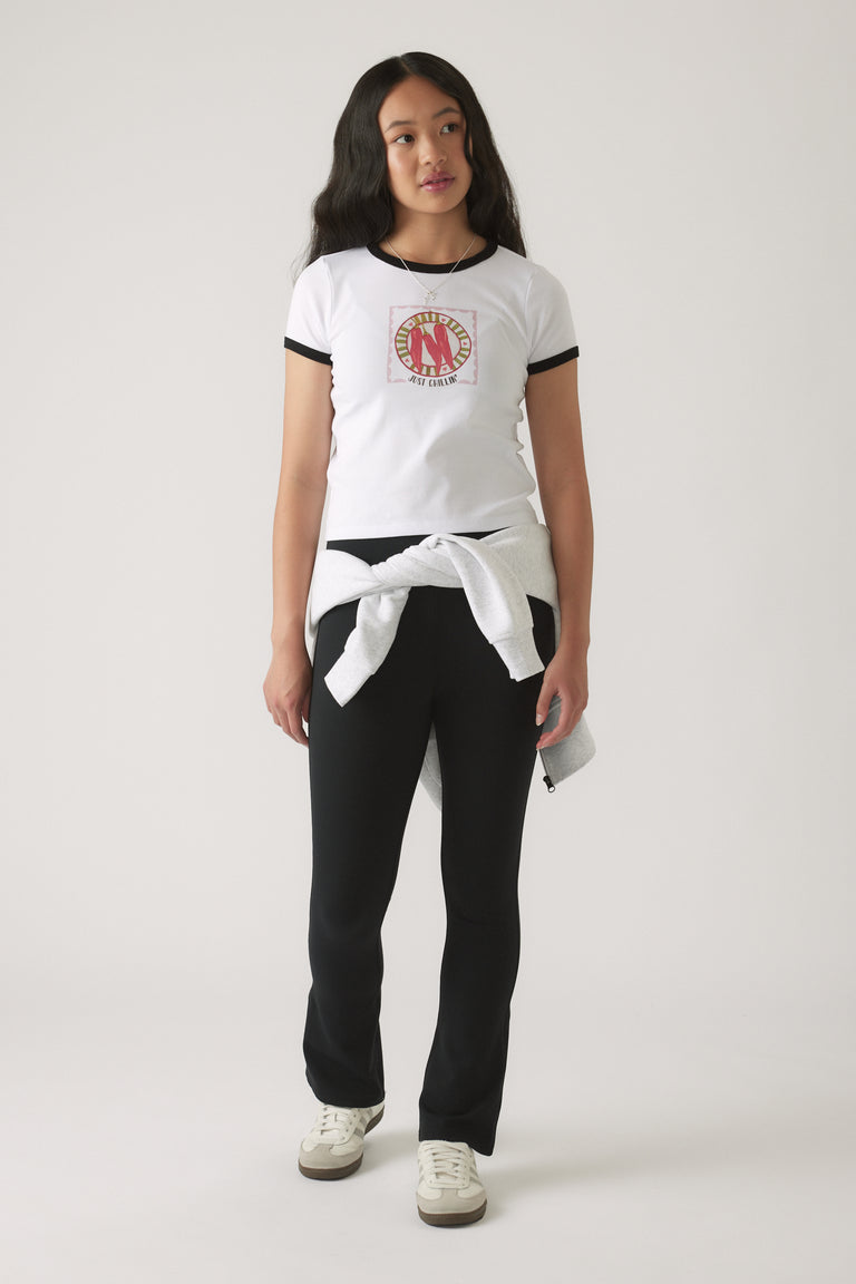 Girl in a white ringer tee with black trim and graphic front, styled with black fitted pants and tied sweatshirt at waist