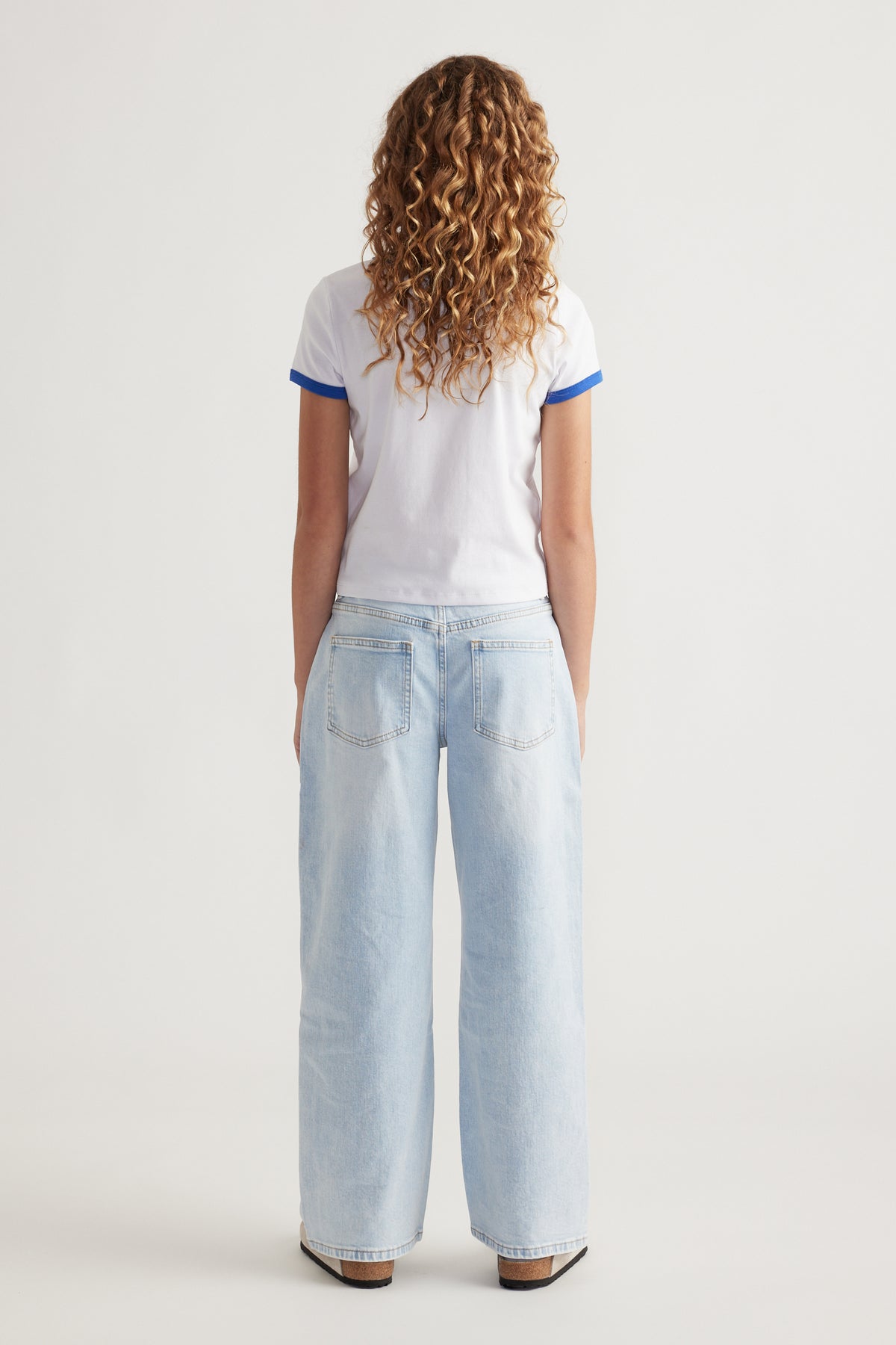 Girl wearing a white cropped tee with blue trim and light wash loose-fitting denim jeans seen from the back