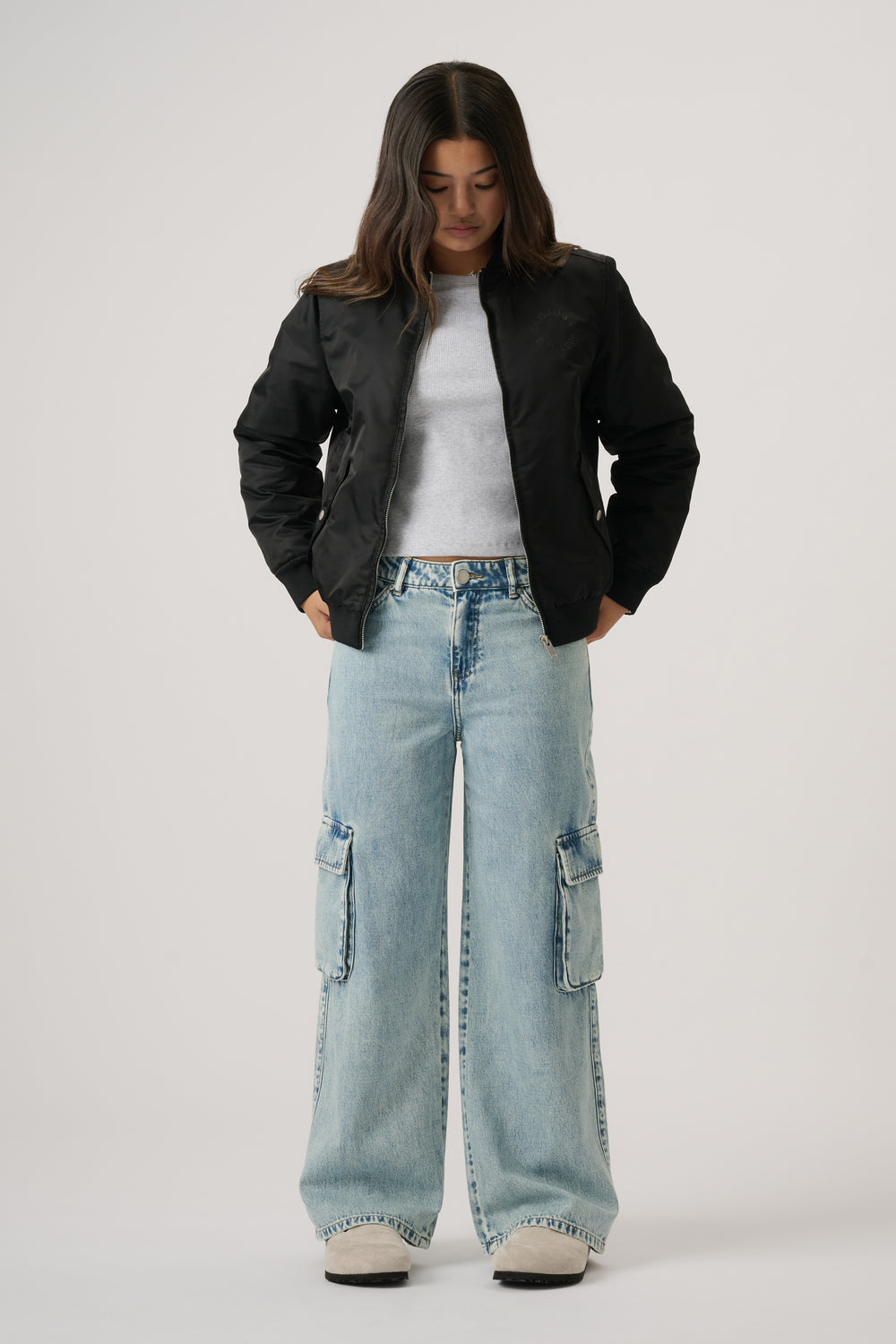 Teen girl wearing light wash denim cargo pants styled with a white tee and black bomber jacket, standing against plain backdrop