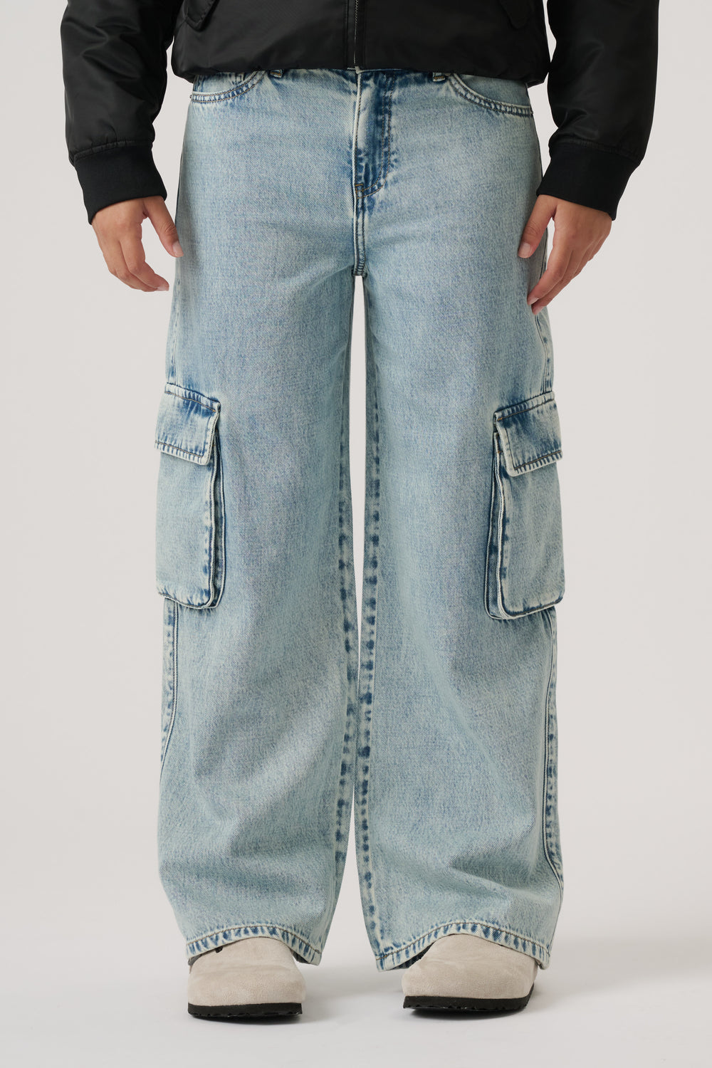 Front view of wide-leg light wash denim cargo pants with large side pockets, styled with a black jacket and white shoes.