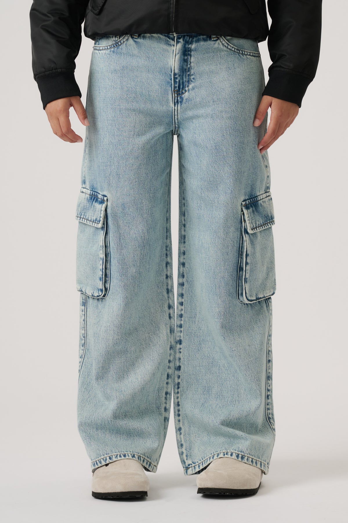 Front view of wide-leg light wash denim cargo pants with large side pockets, styled with a black jacket and white shoes.