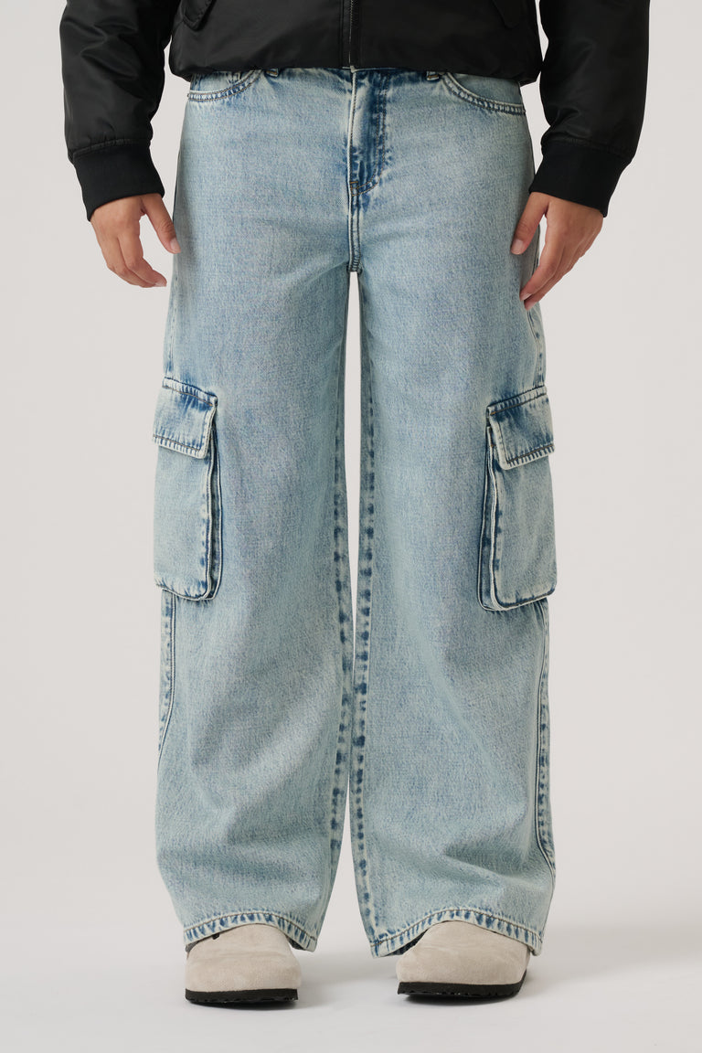Front view of wide-leg light wash denim cargo pants with large side pockets, styled with a black jacket and white shoes.
