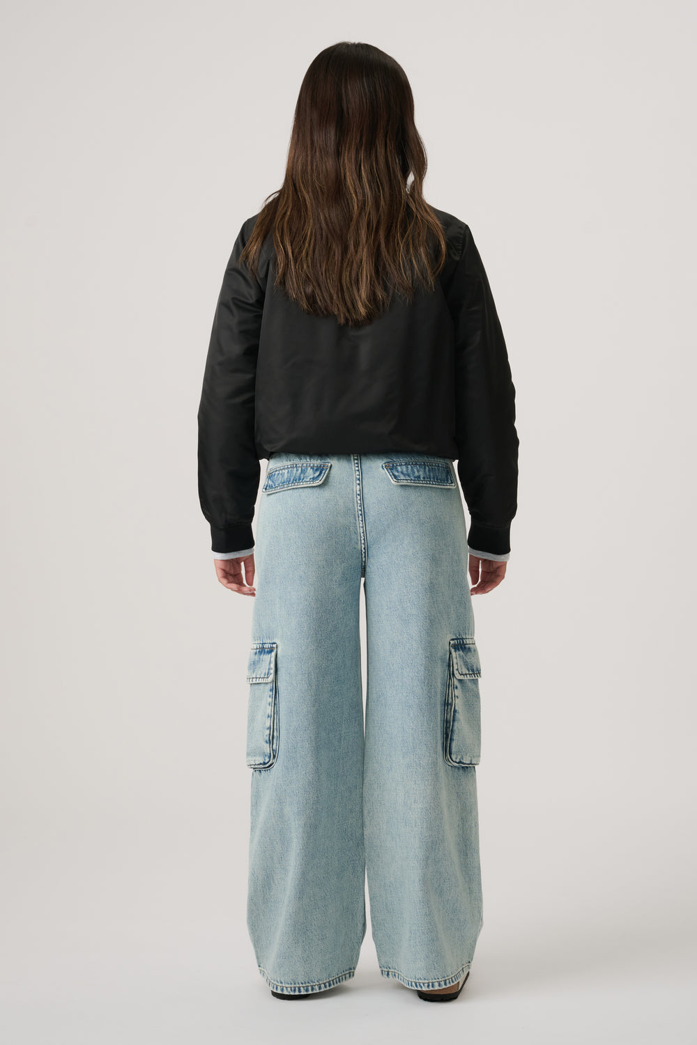 Girl wearing light wash wide-leg denim cargo pants with patch pockets, paired with a black bomber jacket, standing facing away.