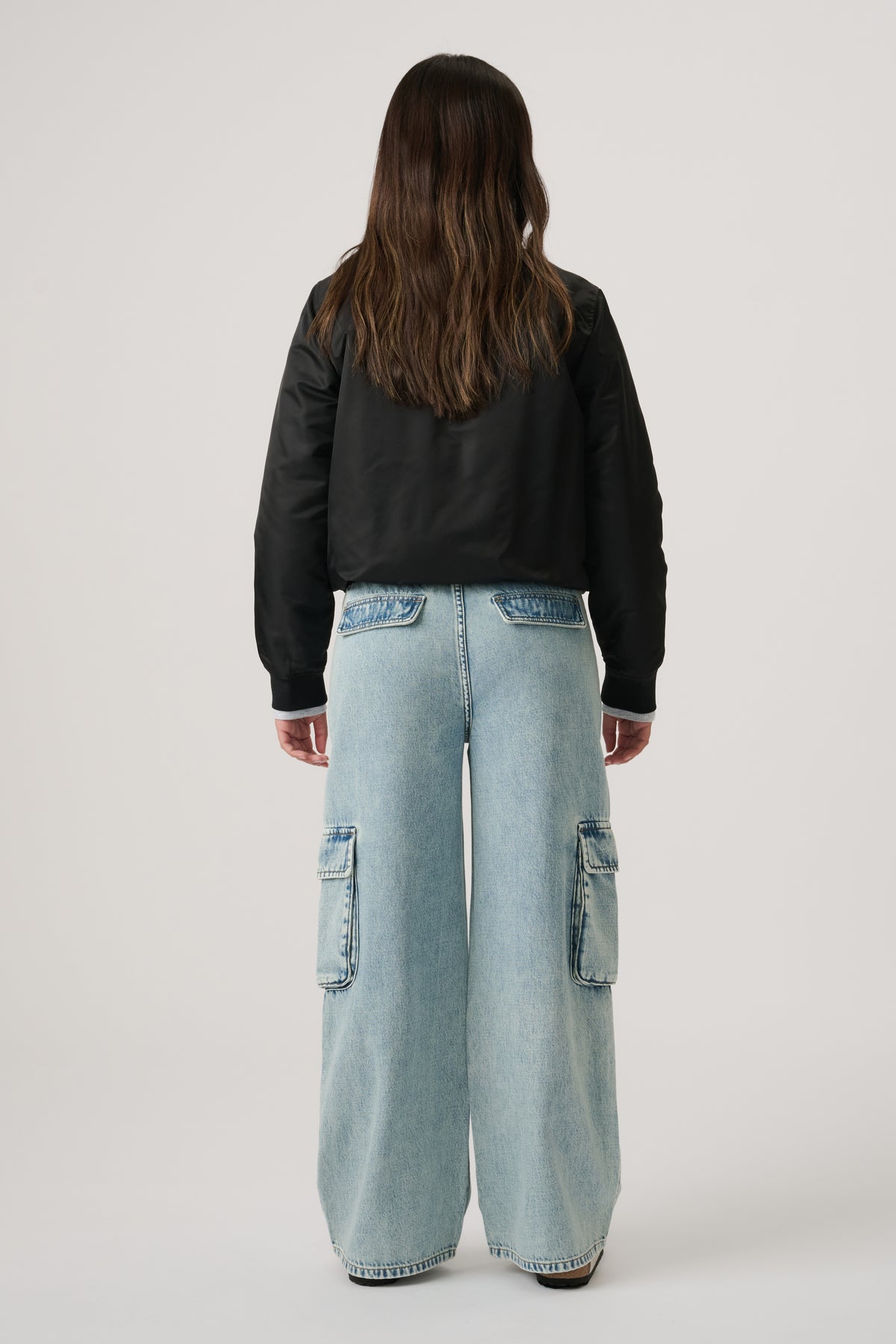 Girl wearing light wash wide-leg denim cargo pants with patch pockets, paired with a black bomber jacket, standing facing away.