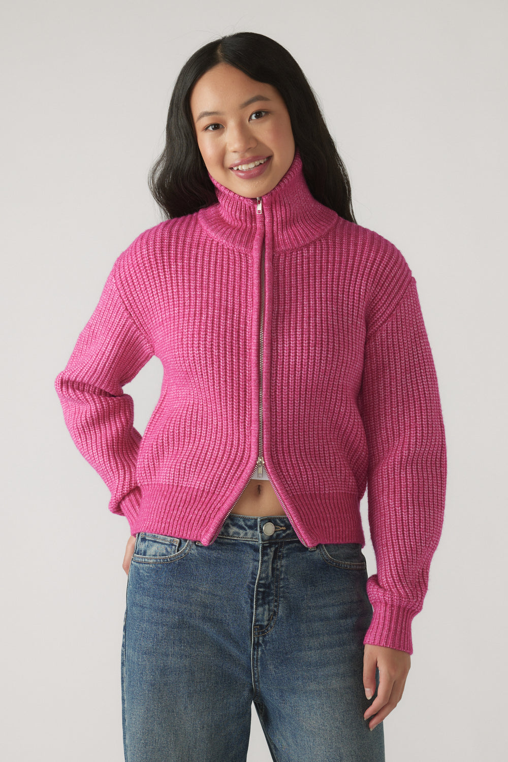 Girl wearing a bright pink ribbed knit zip-up sweater with a high collar paired with relaxed fit blue jeans