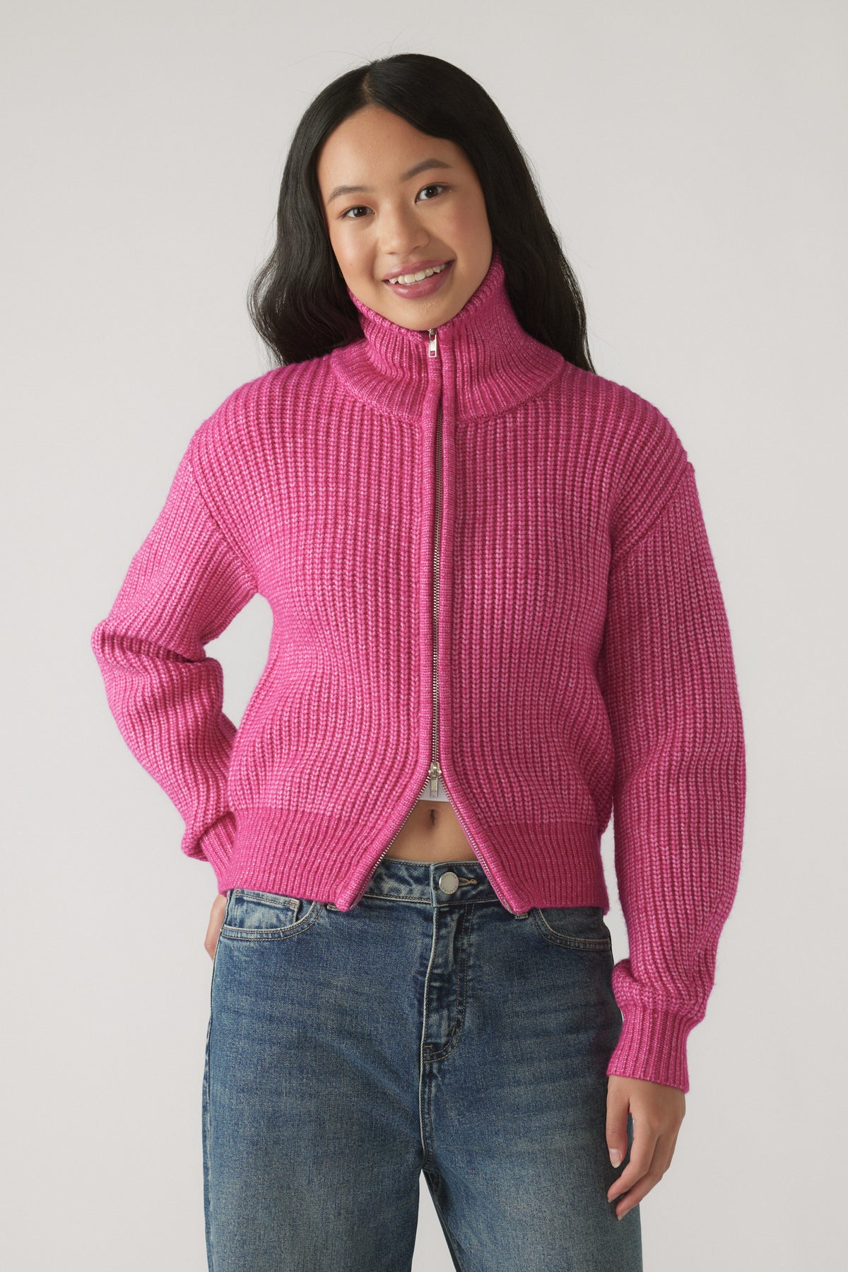 Girl wearing a bright pink ribbed knit zip-up sweater with a high collar paired with relaxed fit blue jeans