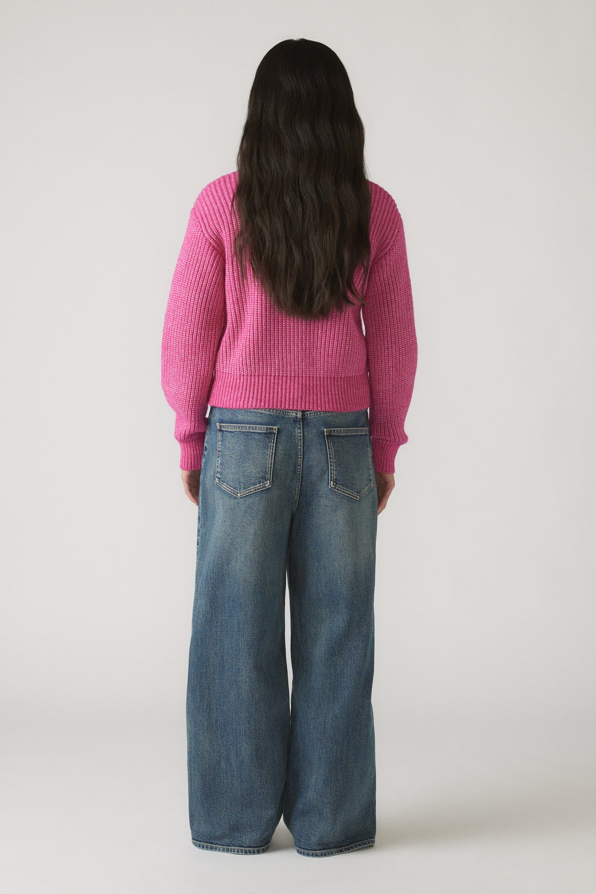 Back view of girl wearing a bright pink knit zip-through sweater paired with loose-fitting blue denim jeans