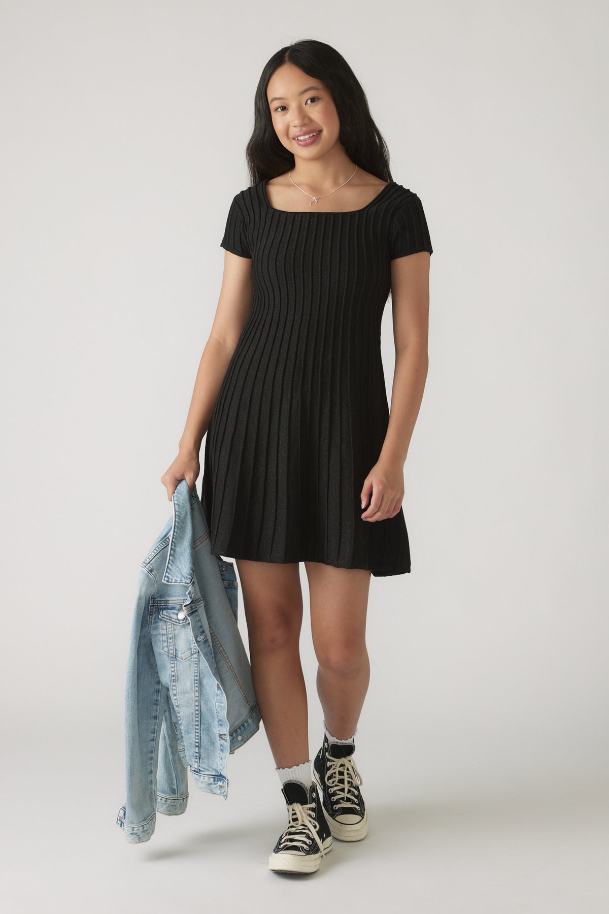 Girl wearing a black Girls Gemma Knit Dress with vertical ribbing, holding a denim jacket, paired with black sneakers and white socks