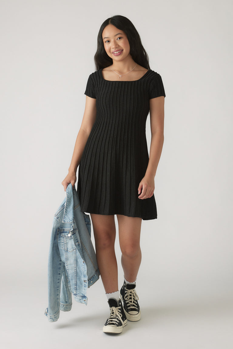 Girl wearing a black Girls Gemma Knit Dress with vertical ribbing, holding a denim jacket, paired with black sneakers and white socks