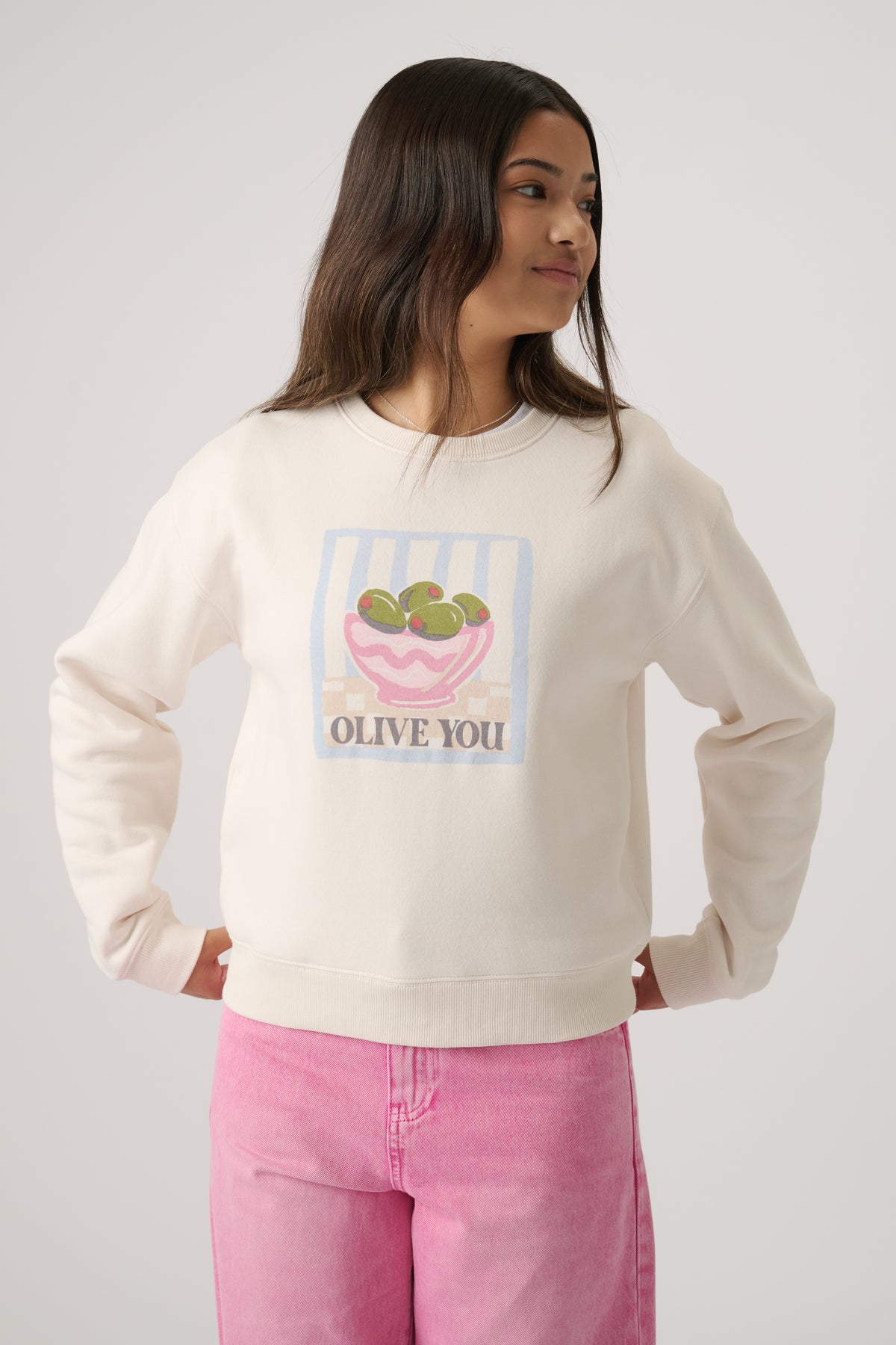 Girl wearing an off-white cropped sweatshirt with a playful olive bowl graphic and light pink jeans in a casual pose