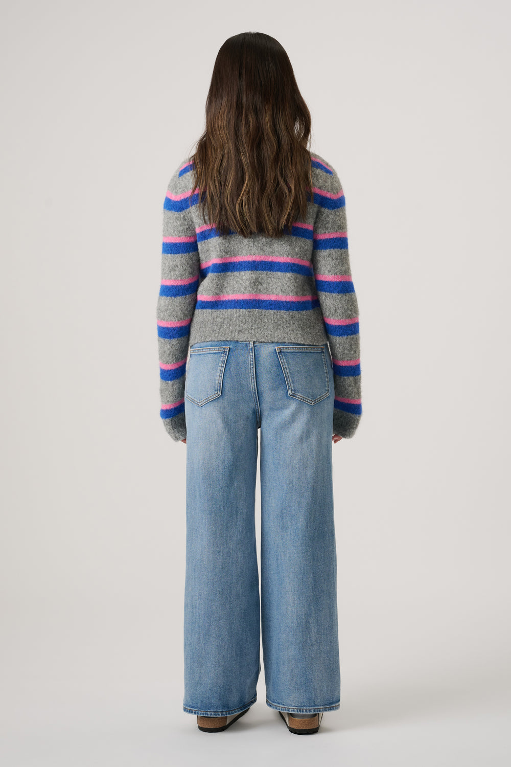 Girl seen from behind wearing a grey cardigan with blue and pink stripes and wide-leg light denim jeans