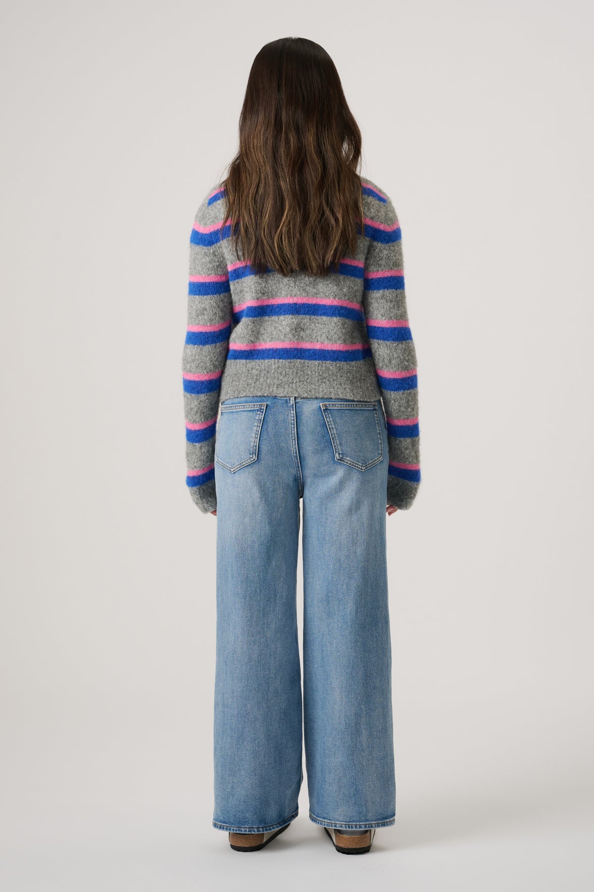 Girl seen from behind wearing a grey cardigan with blue and pink stripes and wide-leg light denim jeans
