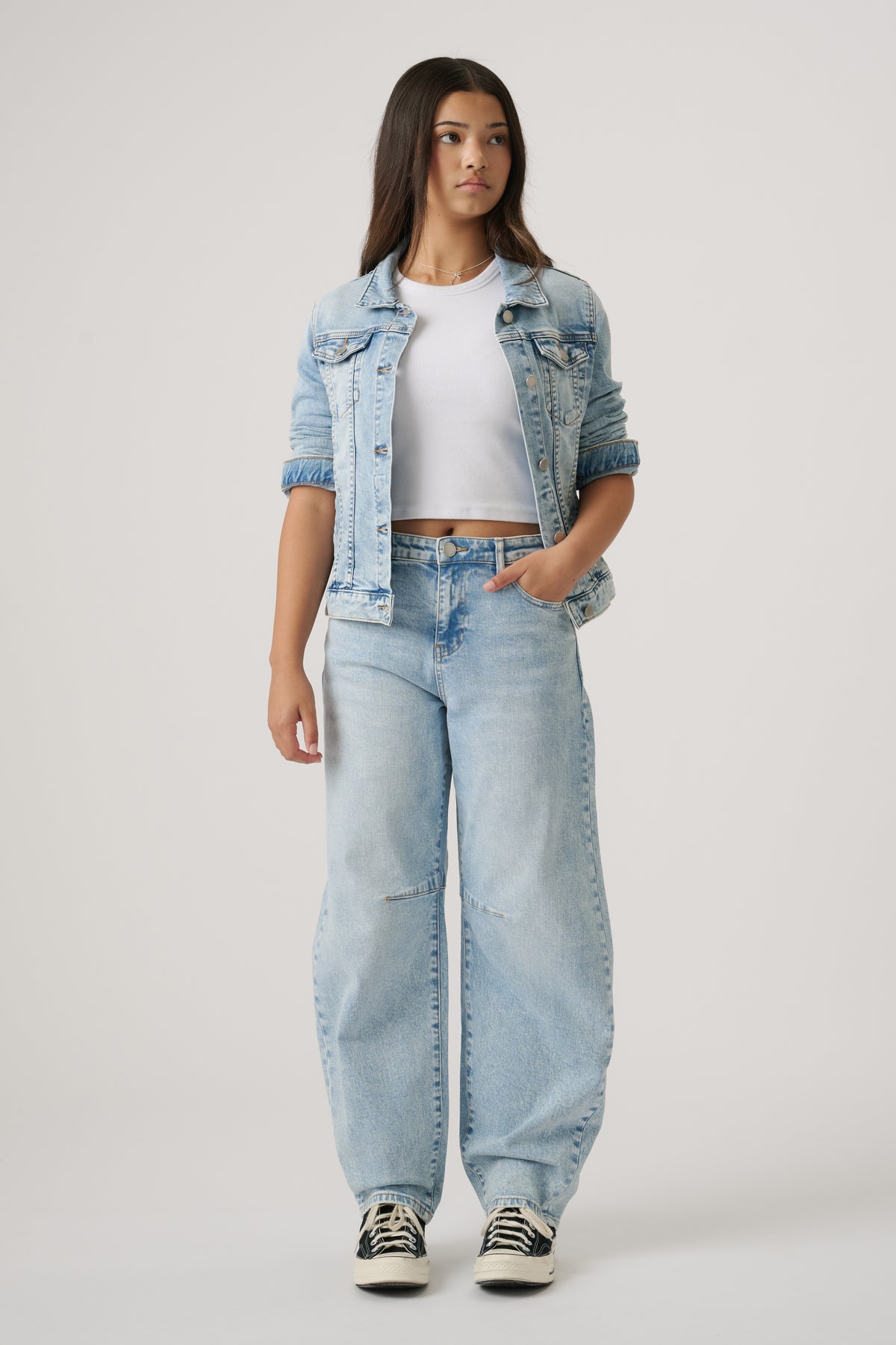 Teen girl wearing light wash denim barrel leg jeans paired with a cropped white top and rolled-up denim jacket