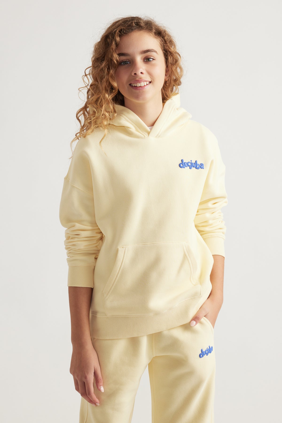 Tween girl smiling wearing a pale yellow oversized hoodie and matching pants with blue logo embroidery