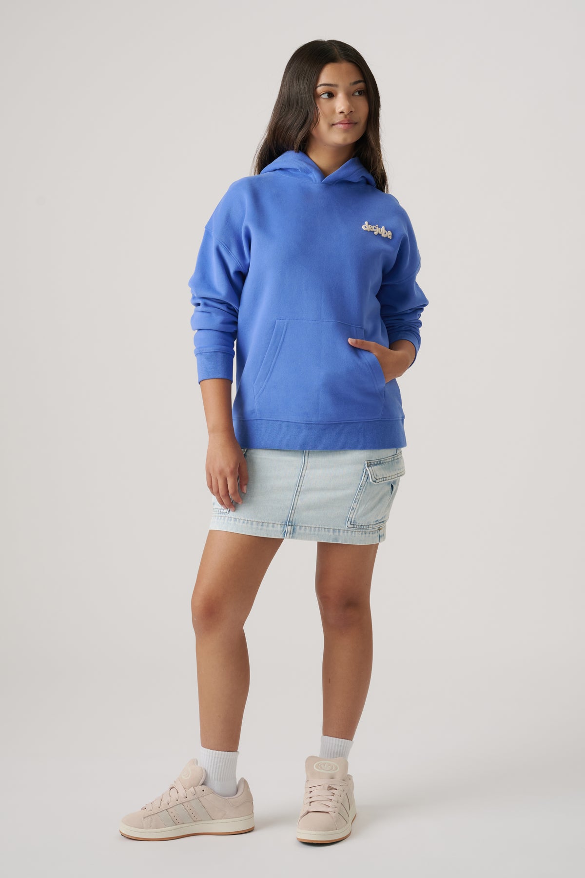 Teen girl wearing a relaxed cornflower blue oversized hoodie paired with a light denim skirt and sneakers.